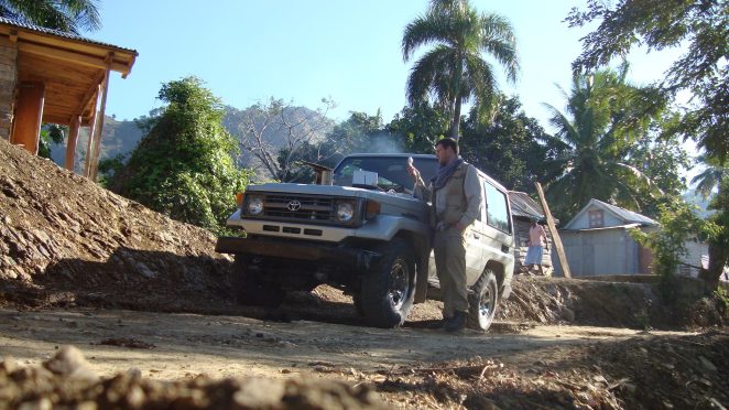A man in outdoor clothing stands next to a Toyota SUV on a dirt path surrounded by tropical trees and a mountainous backdrop.