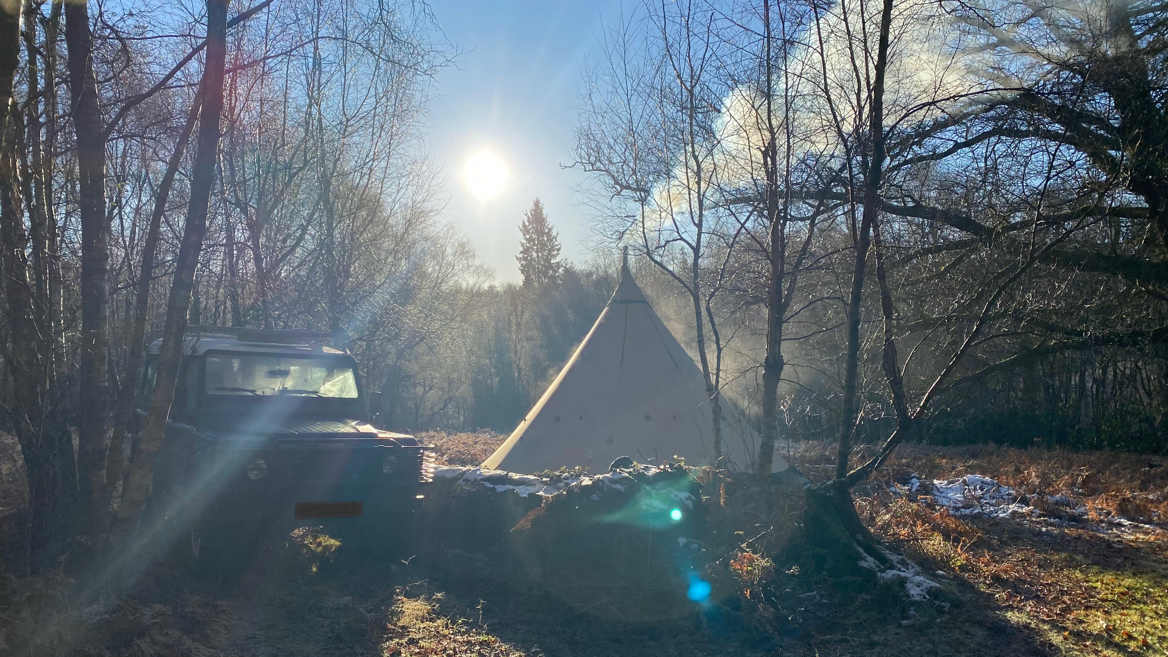 A camping scene featuring a tent and an off-road vehicle surrounded by trees and under clear blue skies, with sunlight shining through branches.
