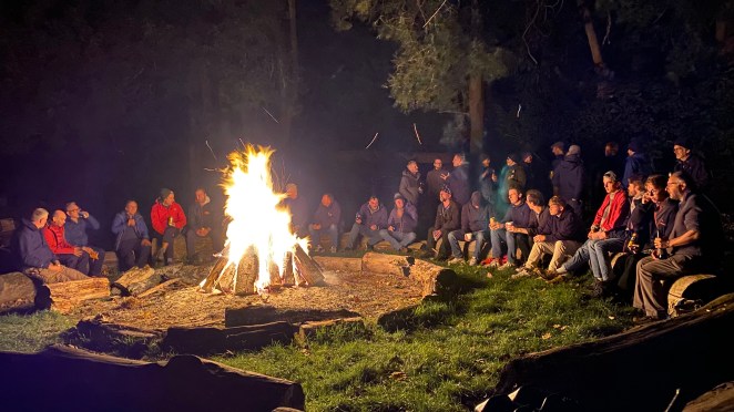A group of people sitting around a large campfire at night, with trees in the background.