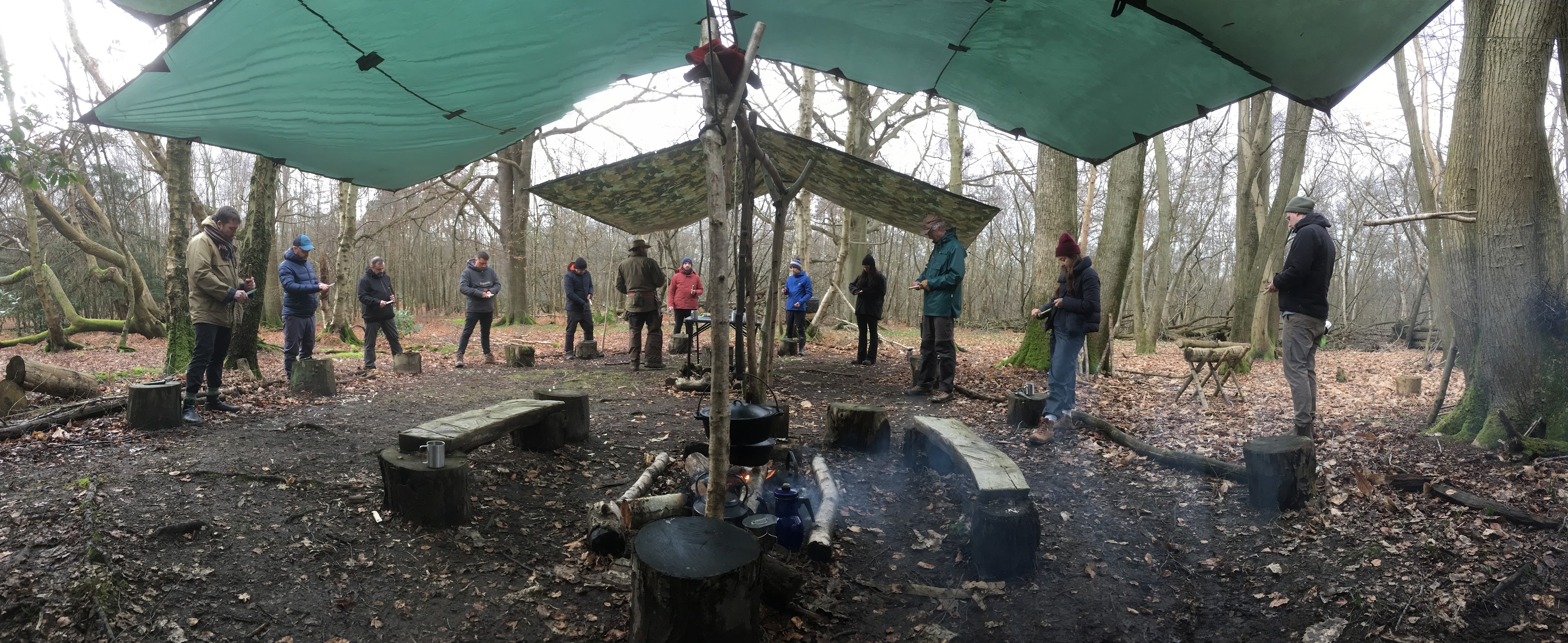 A group of people standing in a wooded area under green tarps, engaging in a workshop or outdoor activity. Stumps and benches are arranged around a fire pit, and there are trees with bare branches in the background.